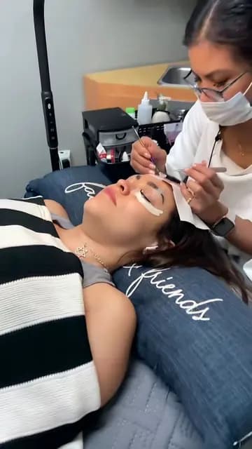 Lash artist in a mask working on a client resting on a striped pillow
